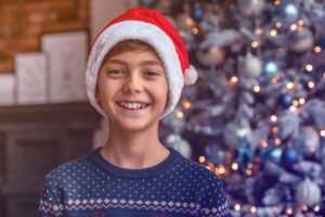 portrait of happy little boy in santa claus hat and standing next to christmas tree in living room