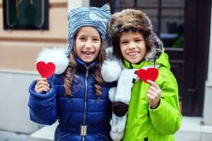 Two smiling children in winter coats and hats hold red heart cutouts. The girl wears a blue coat and knit hat, the boy a green coat and fur hat. Cozy, cheerful scene.