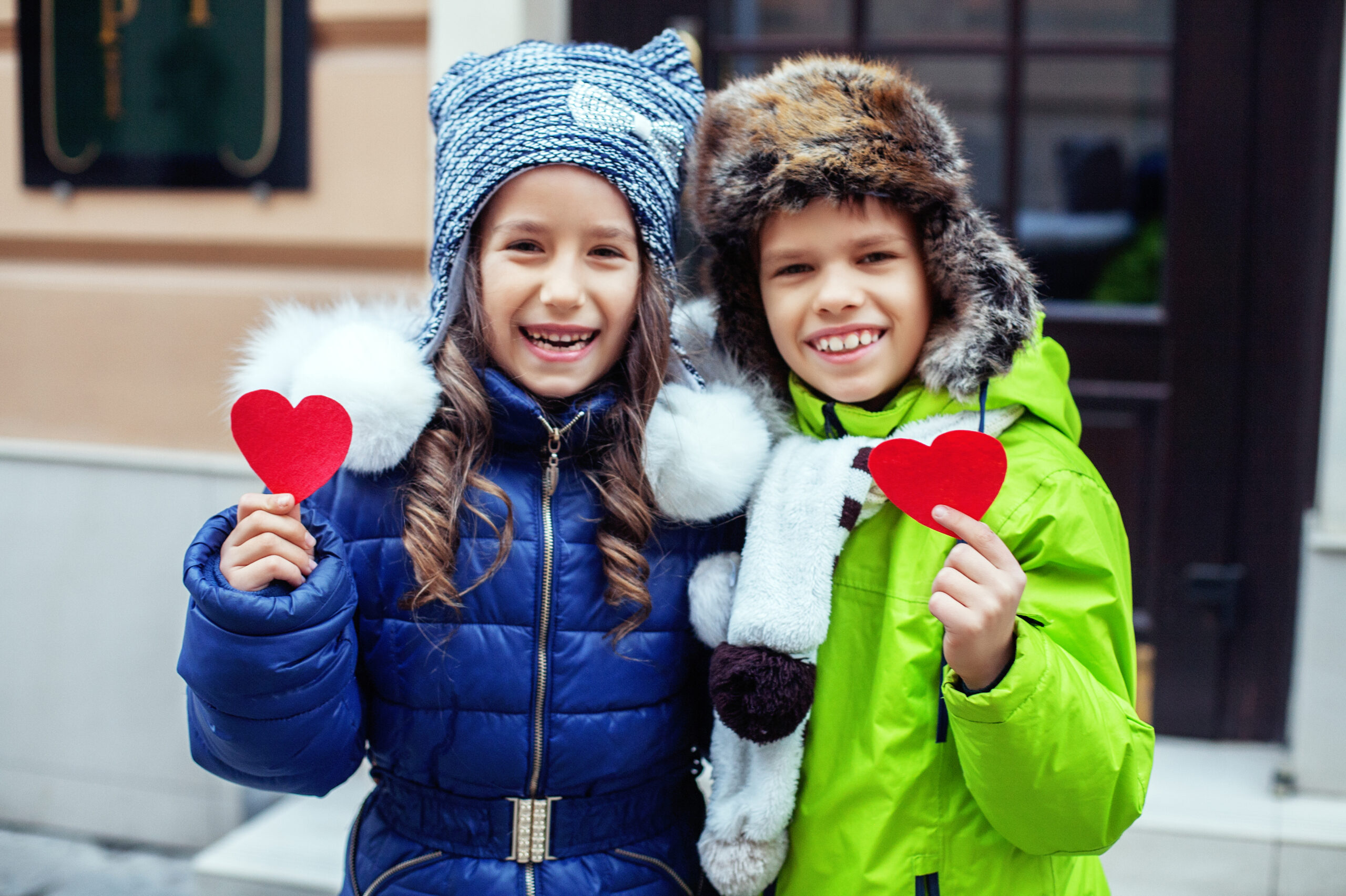 Two smiling children in winter coats and hats hold red heart cutouts. The girl wears a blue coat and knit hat, the boy a green coat and fur hat. Cozy, cheerful scene.