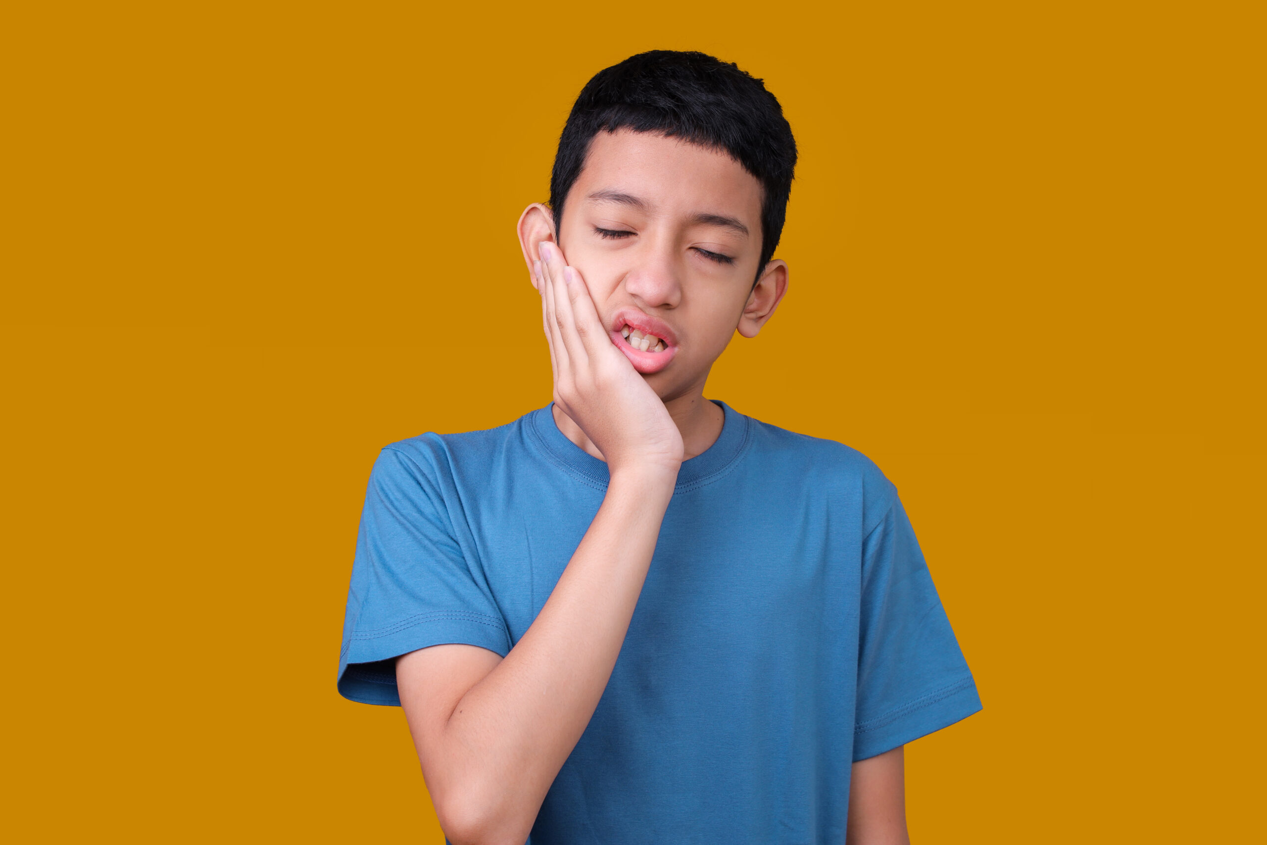 A boy in a blue shirt holds his cheek, grimacing in pain against a mustard yellow background. His expression suggests a toothache.