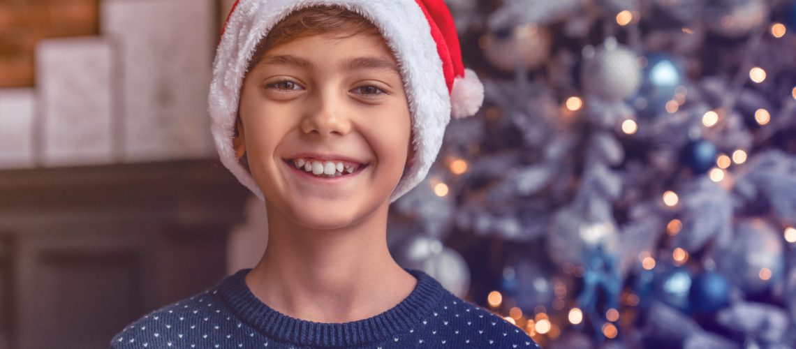 portrait of happy little boy in santa claus hat and standing next to christmas tree in living room