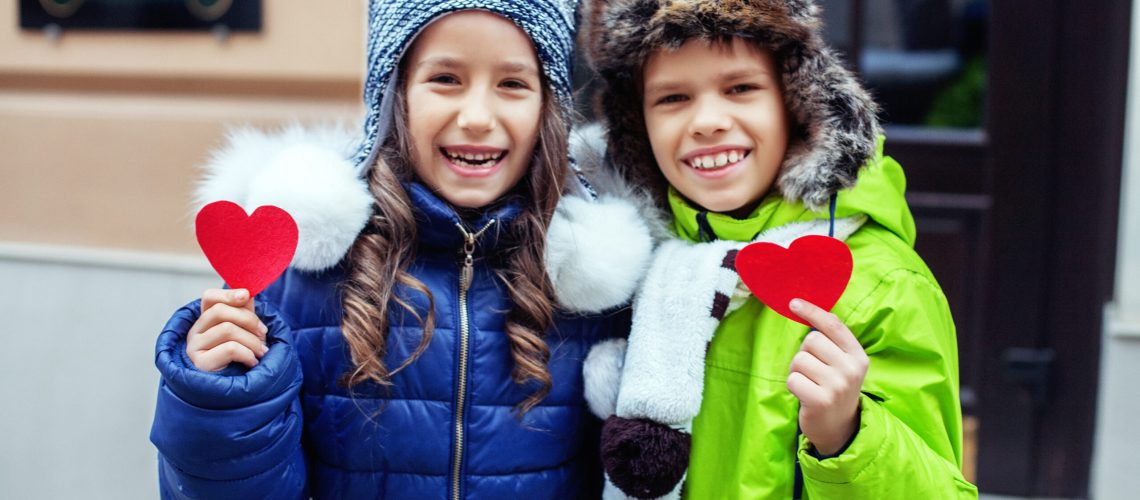 Two smiling children in winter coats and hats hold red heart cutouts. The girl wears a blue coat and knit hat, the boy a green coat and fur hat. Cozy, cheerful scene.