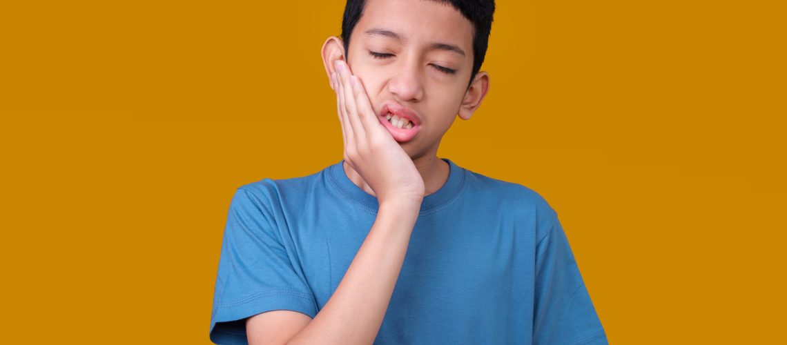 A boy in a blue shirt holds his cheek, grimacing in pain against a mustard yellow background. His expression suggests a toothache.